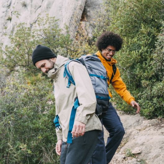 Two men hiking down a rocky trail surrounded by green bushes; one wears a beige jacket and black beanie, the other wears a yellow hoodie and glasses. Both carry backpacks and appear to be enjoying the hike.