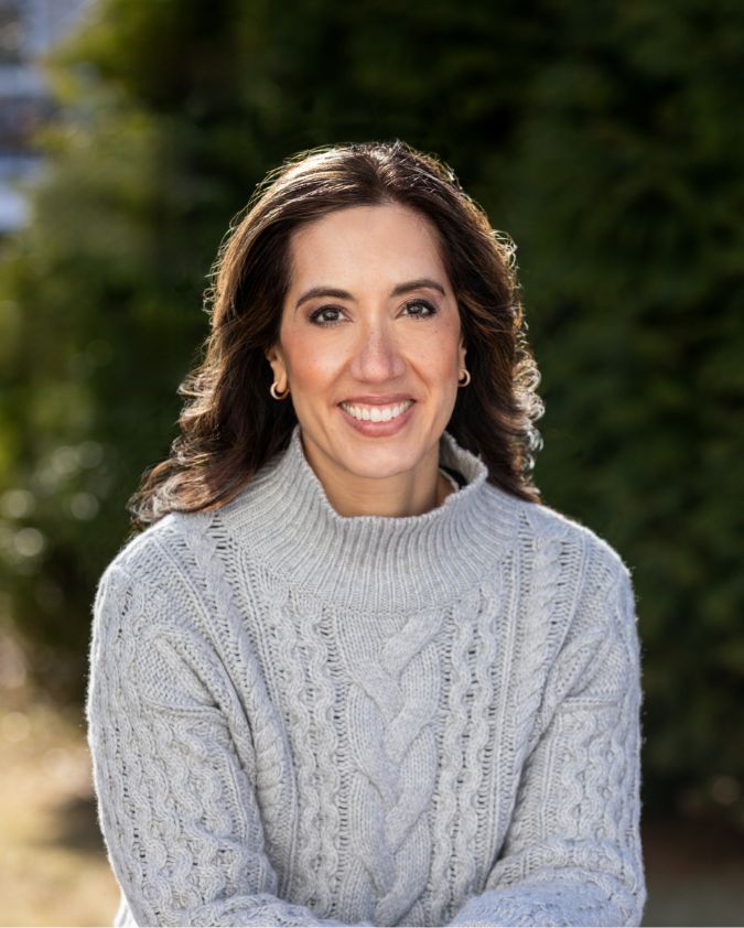 A woman with long brown hair smiles at the camera, wearing a light gray cable-knit sweater, with a blurred green background and sunlight shining on her.