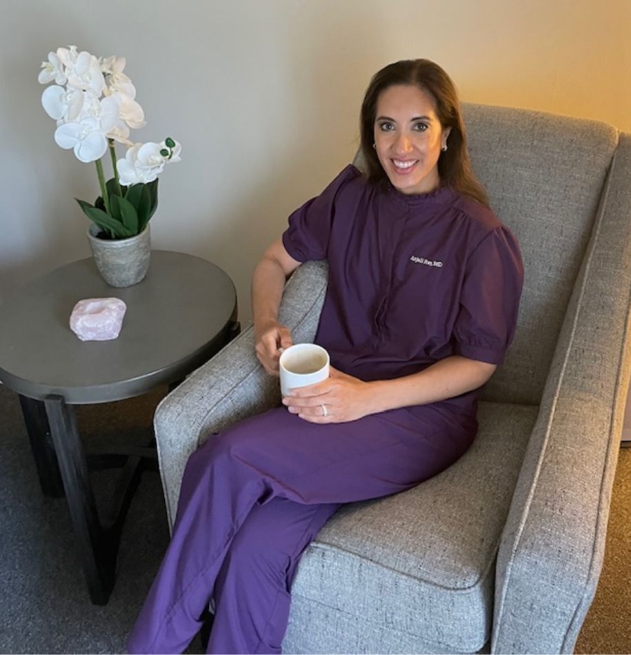 A woman in purple scrubs sits in a gray armchair, smiling and holding a white mug. Next to her is a small table with a potted white orchid and a pink crystal. The setting appears calm and cozy.