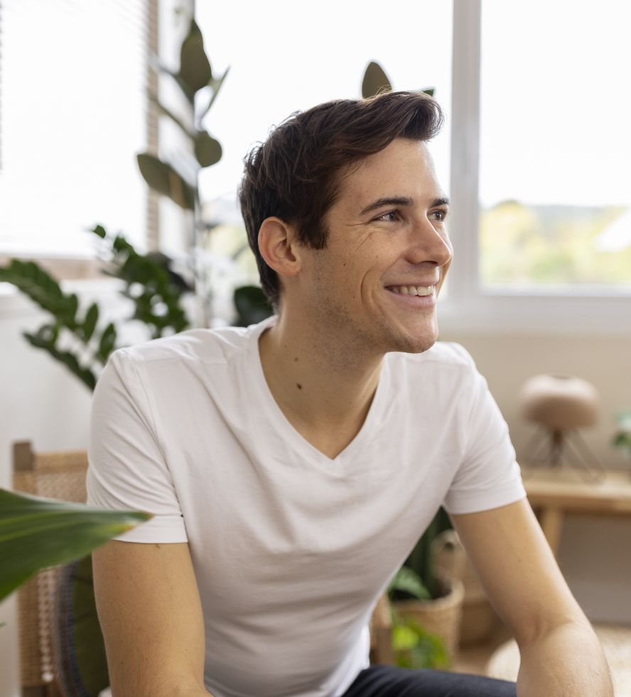 A young man with short brown hair, wearing a white t-shirt, sits indoors smiling and looking to his left. There are green plants and soft natural light in the background.