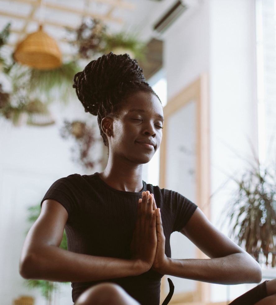 A woman with braided hair sits indoors with her eyes closed, hands pressed together in a prayer position, looking calm and peaceful. She wears a black shirt, surrounded by plants and natural light.