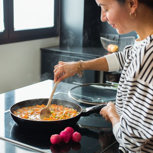 A woman in a striped shirt stirs vegetables in a frying pan on a stovetop, with three radishes nearby. Steam rises from the pan, and sunlight comes through the window in the background.