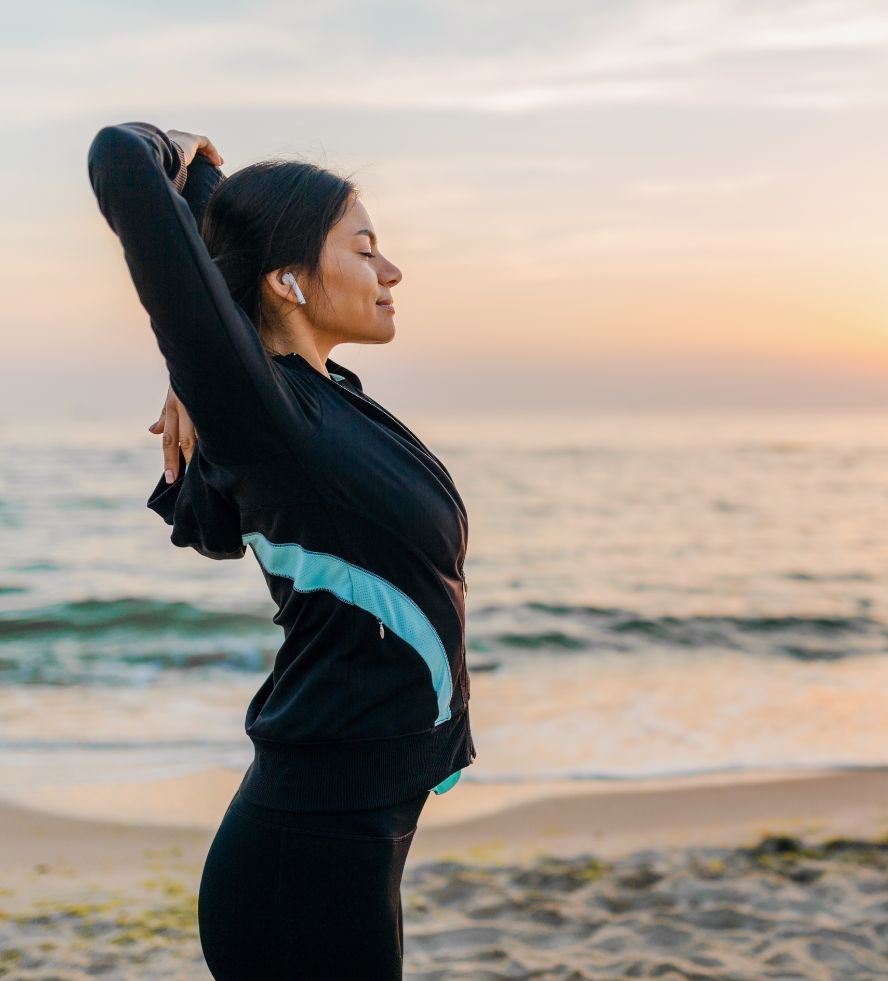 A woman in sportswear stretches her arms behind her head while standing on a beach at sunset, eyes closed and wearing wireless earbuds, with waves and a colorful sky in the background.