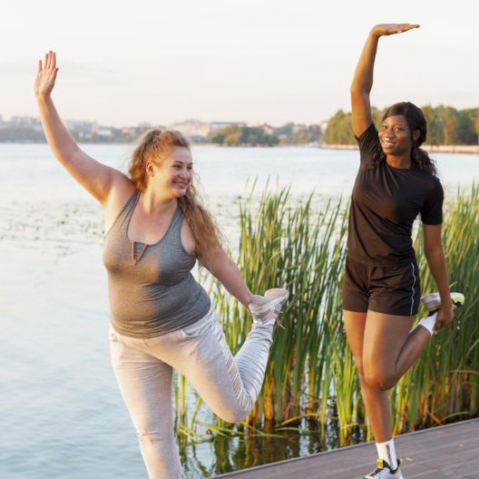 Two women exercising outdoors by a lake, standing on one leg and holding the other foot behind them. Both are smiling and raising one arm overhead, with tall grass and water in the background.