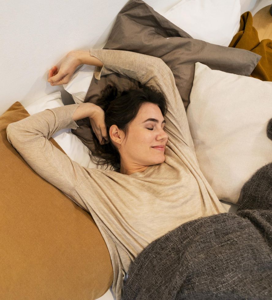 A woman with dark hair is lying in bed, smiling with her eyes closed and stretching her arms above her head. She is covered with a gray blanket and surrounded by white, brown, and beige pillows.
