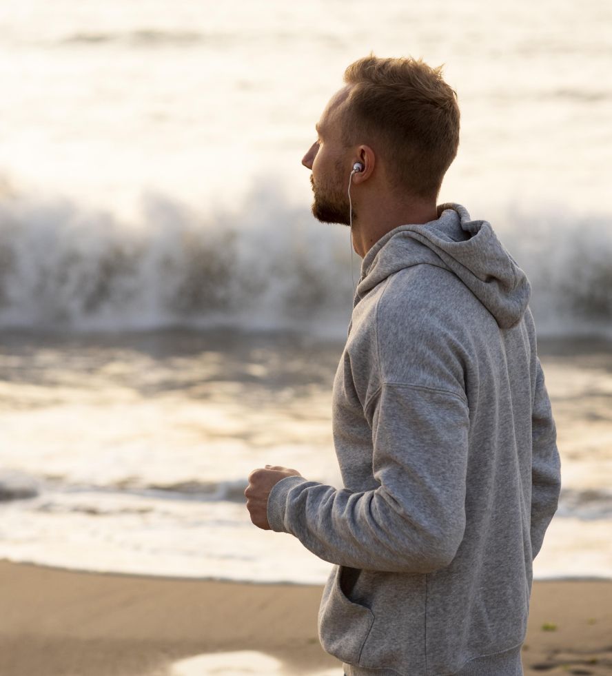 A man in a gray hoodie jogs along the beach at sunset, wearing earphones and looking toward the ocean waves in the background.