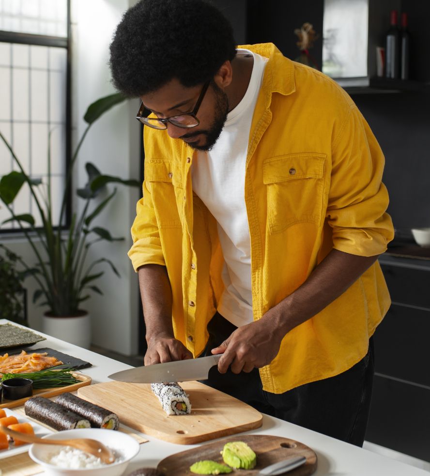 A man wearing a yellow shirt and glasses is slicing a sushi roll on a wooden cutting board in a modern kitchen. Various sushi ingredients and finished rolls are on the counter.