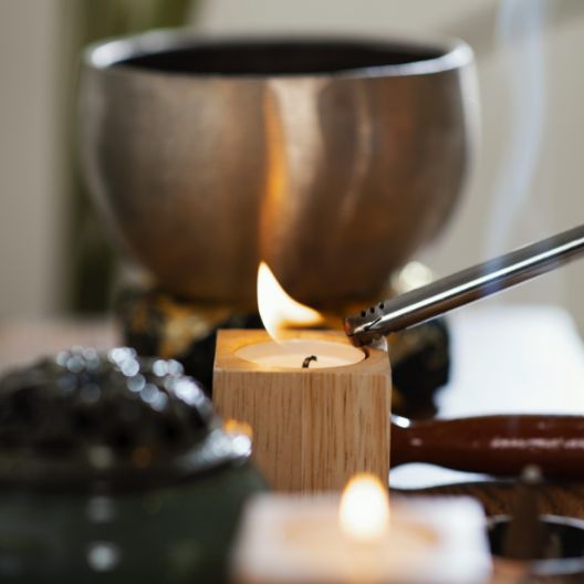 A hand uses a lighter to ignite a small candle in a wooden holder, with a metal bowl and another lit candle blurred in the background.