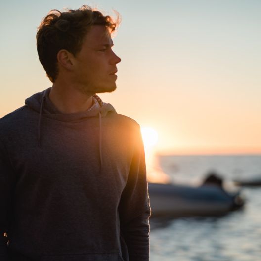 A man wearing a dark hoodie stands near the water at sunset, looking to his left. The sun is low on the horizon, casting a warm glow and creating a silhouette effect on his profile.